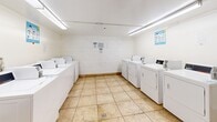 Located at 1250 N June St in Los Angeles, this shared laundry facility is a single-room space designed to serve the practical needs of building residents. The room is laid out efficiently, housing a generous bank of commercial-grade top-loading washing machines along one wall and a row of standalone dryers along the opposite side, providing ample capacity for multiple users at once. The space is finished with durable, easy-to-clean tan tile flooring throughout, and the white-painted walls and cinder block accents give the room a clean, utilitarian appearance. Overhead fluorescent lighting ensures the space is well-lit at all times. Two folding tables are positioned within the room, offering convenient surfaces for sorting and folding laundry. Informational signage is posted on the walls, including usage guidelines and etiquette reminders for shared use. The layout allows for comfortable movement between machines and folding areas. Overall, this laundry facility is a functional, well-equipped communal space designed with practicality and ease of use in mind, serving as a valuable shared amenity for residents of the property.