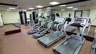 This fitness center offers a thoroughly equipped workout environment designed to accommodate a wide variety of exercise routines. The space is outfitted with multiple treadmills and elliptical cross-trainers positioned along the floor, providing ample cardio options for users of all fitness levels. Complementing the cardio section is a dedicated strength-training area featuring a selection of cable and resistance machines, allowing for targeted muscle group workouts. The floor is covered in durable rubberized cork-style material, which provides cushioning and stability underfoot — a practical and safety-conscious choice for a high-traffic fitness environment. Full-length mirrors line the walls, visually expanding the space and allowing users to monitor their form during exercise. Overhead, a grid of recessed and panel lighting ensures the room is consistently and evenly illuminated throughout. The layout is organized and spacious, with equipment arranged to allow comfortable movement between stations. The overall atmosphere is functional and professional, making this fitness center a practical and well-maintained facility suited for daily use.