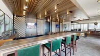 Located in the heart of Houston's vibrant Montrose neighborhood, this upscale amenity space delivers a refined, contemporary experience across a generously sized single-floor layout. The interior design blends bold textures and rich materials throughout, including striated wood-paneled ceilings, polished large-format tile flooring, and striking perforated decorative wall panels that add depth and visual interest.

The main lounge area features an elegant seating arrangement with tufted leather sofas, accent chairs, and curated artwork, all beneath a statement ceiling detail inlaid with a dramatic marbled panel. Recessed and pendant lighting—including eye-catching metallic globe fixtures—creates a warm, inviting ambiance. A dedicated bar area showcases a long counter with green velvet bar seating, wood-grain striped ceiling treatments, and multiple mounted displays, ideal for social gatherings.

A separate lounge zone with a billiards table and additional leather seating offers a more relaxed setting, while a kitchen and dining nook with iridescent tile backsplash and pendant lighting serves as a refined entertaining hub. Floor-to-ceiling sliding glass doors open to multiple wraparound outdoor terraces with glass-railed balconies, warm wood-clad ceilings, and sweeping views of the Houston skyline—making this space as impressive outside as it is within.