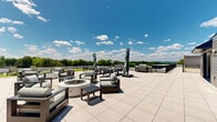 Located at 144 N Main St in Dublin, PA, this rooftop amenity space offers a well-appointed combination of indoor and outdoor areas designed for relaxation and entertainment. The interior bar area features a granite-topped island, sleek blue cabinetry with gold fixtures, a built-in wine cooler, and a striking green tile backsplash with open wood shelving. A glass-panel garage door opens fully to seamlessly connect the interior to the expansive outdoor terrace, blurring the boundary between inside and out. The wide-format tile terrace extends generously across the rooftop, providing ample room for seating and gathering. A wood-clad pergola anchors one section of the outdoor space, complete with string lighting and a mounted outdoor television, creating a cozy al fresco lounge area. A separate section of the terrace features a lush green lawn area alongside additional seating arrangements. The perimeter of the rooftop is lined with sturdy metal railings, offering unobstructed panoramic views of the lush, tree-lined landscape and open sky beyond. Modern wall-mounted light fixtures and clean horizontal siding complete the refined exterior aesthetic of this elevated outdoor retreat.