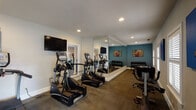 This fitness center in Mobile, Alabama, offers a thoughtfully designed single-floor workout facility built to support a well-rounded exercise routine. The space features a clean, open layout with a neutral color palette accented by a bold teal feature wall, creating an energizing yet focused atmosphere throughout.

The cardio zone is well-stocked with multiple True-brand elliptical trainers, stair climbers, and treadmills arranged across durable, carpet-tiled flooring. Wall-mounted flat-screen TVs are positioned for easy viewing during workouts, adding a convenient entertainment element to the space.

A large floor-to-ceiling mirror wall expands the sense of space and allows users to monitor their form during exercise. Adjacent to the mirror wall, a dedicated free weights area houses a full rack of dumbbells alongside a workout bench, making it easy to transition between strength and cardio training.

Natural light filters in through shuttered windows along one wall, while recessed ceiling lighting ensures consistent illumination throughout. A set of French doors with grid-pane windows provides an exit to an outdoor area, offering views of the surrounding grounds and bringing an airy, open feel to the interior. Vibrant fitness-themed artwork displayed on the teal walls adds a touch of personality to the functional space.