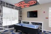 This dynamic, single-floor game room is designed to deliver an immersive entertainment experience through bold design choices and a diverse lineup of recreational amenities. The space is defined by its striking geometric-patterned carpet in blue, beige, and brown tones, which anchors the room's energetic aesthetic. Vivid orange accent walls contrast with gray and neutral tones throughout, creating a lively and engaging atmosphere.

At the heart of the room sits a full-size air hockey table, accompanied by a wall-mounted flat-screen television and a distinctive neon orange sculptural chandelier that serves as a standout focal point overhead. A city map-inspired mural wallpaper adds an urban, graphic edge to one wall, reinforcing the room's contemporary design language.

The space also features multiple upright arcade machines positioned along the walls, offering classic gaming entertainment. A unique lounge nook showcases high-back booth seating adorned with graffiti-style artwork, paired with yellow pendant lighting and glass-top side tables — providing a stylish spot to relax between games. Teal stools and a bar-height counter area offer additional casual seating options.

Natural light filters in through several windows fitted with blinds, while recessed ceiling lighting and a wall-mounted mini-split unit ensure year-round comfort. The overall ambiance is bold, urban, and purpose-built for social recreation.