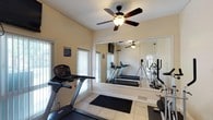 This single-room fitness center is designed to provide a functional and comfortable workout environment. The space is finished with light-toned walls and neutral ceramic tile flooring, creating a clean and bright atmosphere. A large wall-to-wall mirror on one side of the room visually expands the space and allows users to monitor their form during exercise. The room is equipped with multiple treadmills and a stationary cycle, offering a variety of cardio workout options. A wall-mounted flat-screen television is positioned near the windows, providing entertainment during workouts. Natural light filters in through several windows fitted with blinds, adding to the open and airy feel of the space. A ceiling fan with an integrated light fixture provides air circulation and additional illumination, keeping the environment comfortable during use. A French door with glass panes connects to an adjacent area, enhancing the sense of openness. Rubber floor mats are placed beneath the equipment for safety and floor protection. The textured ceiling and straightforward layout contribute to a practical, no-frills fitness facility well-suited for daily use.