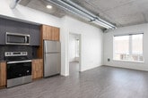 This thoughtfully designed unit blends modern finishes with bold industrial character throughout its single-floor layout. Exposed concrete ceilings with visible ductwork and structural beams set a striking architectural tone, while wood-grain vinyl flooring runs consistently across the living areas, bedrooms, and kitchen, unifying the space with warmth and texture.

The open-concept living and kitchen area is generously proportioned, allowing ample natural light to pour in through large windows. The kitchen is a standout feature, outfitted with sleek two-tone cabinetry in dark charcoal and natural wood-grain finishes, a dark tile subway backsplash, stainless steel appliances including a refrigerator, range, microwave, and dishwasher, and a single-basin sink.

The bedrooms offer a clean, minimalist aesthetic with the same exposed concrete ceilings and wood-grain flooring. One bedroom features direct access to a private outdoor space through a sliding glass door, framing views of the surrounding neighborhood. Natural light is a consistent feature across all rooms.

The bathroom is finished with a clean, contemporary palette, featuring a tub-shower combination with a curved rod, a wood-toned vanity with a white countertop, and a large mirror. The overall ambiance of the space is urban and modern, with industrial architectural elements balanced by clean white walls and warm wood tones.