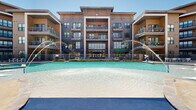 Located at 2401 Linden Dr in Denton, Texas, this expansive outdoor amenity area is designed to deliver a resort-style experience within a modern residential community. At its centerpiece is a generously sized swimming pool featuring elegant water fountains that arc gracefully across the surface, complemented by a separate spa and a shallow beach-entry section. The pool deck is finished in smooth, light-toned concrete and is lined with contemporary white lounge chairs and seating arrangements, ideal for relaxing under the Texas sun.

A covered cabana area anchors one side of the deck, framed by substantial natural stone columns and equipped with ceiling fans, built-in speakers, wall-mounted screens, and stylish outdoor furniture arranged around a central coffee table — creating a comfortable shaded retreat. Additional pergola structures provide further shade along the perimeter.

Adjacent courtyard spaces feature artificial turf areas, string lighting, and modern white modular seating set against warm wood-clad exterior walls and expansive floor-to-ceiling glass. Ornamental landscaping with native grasses, trimmed hedges, and decorative rock borders adds to the polished aesthetic. Iron fencing with gated pool access ensures security throughout. The multi-story residential buildings surrounding the amenity area create an intimate, enclosed atmosphere while overlooking the entire outdoor space.