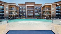 Located at 2401 Linden Dr in Denton, Texas, this expansive outdoor amenity area is designed to deliver a resort-style experience within a modern residential community. At its centerpiece is a generously sized swimming pool featuring elegant water fountains that arc gracefully across the surface, complemented by a separate spa and a shallow beach-entry section. The pool deck is finished in smooth, light-toned concrete and is lined with contemporary white lounge chairs and seating arrangements, ideal for relaxing under the Texas sun.

A covered cabana area anchors one side of the deck, framed by substantial natural stone columns and equipped with ceiling fans, built-in speakers, wall-mounted screens, and stylish outdoor furniture arranged around a central coffee table — creating a comfortable shaded retreat. Additional pergola structures provide further shade along the perimeter.

Adjacent courtyard spaces feature artificial turf areas, string lighting, and modern white modular seating set against warm wood-clad exterior walls and expansive floor-to-ceiling glass. Ornamental landscaping with native grasses, trimmed hedges, and decorative rock borders adds to the polished aesthetic. Iron fencing with gated pool access ensures security throughout. The multi-story residential buildings surrounding the amenity area create an intimate, enclosed atmosphere while overlooking the entire outdoor space.