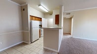 This well-organized single-floor, one-bedroom, one-bathroom unit offers a practical and comfortable layout across a compact but functional footprint. The open-concept living area seamlessly combines the kitchen, dining, and living spaces, all finished with light tile flooring. The kitchen features warm wood-toned cabinetry, white appliances including a refrigerator and range, and a peninsula with a laminate countertop that doubles as a breakfast bar and natural room divider. Crown molding accents the dining and living area, adding a refined touch to the neutral, beige-toned interior. The living area is carpeted, bright, and includes a ceiling fan, with windows fitted with blinds allowing natural light to filter in. A dedicated pantry provides additional kitchen storage, while a separate in-unit laundry nook with washer/dryer hookups adds everyday convenience. The bedroom is generously proportioned, carpeted, and brightened by multiple windows with outdoor views. It benefits from crown molding, a ceiling fan, and access to a spacious walk-in closet with built-in shelving and hanging rods. An additional closet provides further storage. The bathroom is clean and functional, featuring a full tub-shower combo with white tile surround, a toilet, and tile flooring. A hallway connects the private areas of the unit efficiently.
