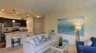 Located at 130 Ocean Aisle Circle in Salisbury, Maryland, this single-floor residence offers a thoughtfully designed open-concept layout with a warm, coastal aesthetic throughout. The living area serves as the heart of the home, featuring plush carpeting underfoot and neutral-toned walls that create a bright, inviting atmosphere. A large ocean-inspired wall mural acts as a striking focal point, reinforcing the coastal theme and adding a sense of depth to the space. The living room is comfortably appointed with ample seating and a glass-top coffee table, making it ideal for both relaxing and entertaining. The kitchen flows seamlessly from the living area, separated by a breakfast bar with seating — a practical and social feature for everyday living. Dark cabinetry contrasts elegantly with light granite countertops, while pendant lighting adds a stylish touch above the bar. Stainless steel appliances, including a range and microwave, complete the well-equipped kitchen. Recessed lighting throughout the kitchen ensures the space is both functional and visually appealing. The entry door is conveniently accessible from the main living space, contributing to the home's efficient and cohesive single-level floor plan. Overall, the space exudes a relaxed, beach-inspired charm.