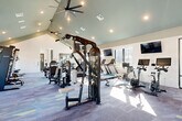 Located at The Pullman in Union City, Georgia, this expansive fitness center offers a generously proportioned, single-floor layout designed to support a full spectrum of workout routines. The space is immediately distinguished by its soaring vaulted ceiling, which rises dramatically along the central ridge and is complemented by a teal-painted upper accent, recessed lighting, and large ceiling fans that maintain comfortable airflow throughout. Natural light pours in through numerous windows lining the perimeter walls, with views of the surrounding outdoor amenities, including what appears to be a pool area, creating an energizing atmosphere. The entry foyer features a branded "The Pullman" welcome mat, built-in shelving, and a sleek wood bench, providing a clean and organized transition into the main floor. The fitness floor itself is carpeted in a geometric, multi-toned pattern and is comprehensively outfitted with cable machines, selectorized strength stations, a Smith machine, squat rack, free weight benches, a full dumbbell rack, kettlebells, medicine balls, resistance training accessories, treadmills, stair climbers, stationary bikes, and rowing machines. Wall-mounted flat-screen televisions are positioned throughout for entertainment during workouts. Full-length mirrors enhance the sense of openness while serving functional purposes for form checking.