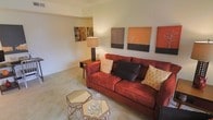 This single-floor living space centers around a inviting living room designed with a warm, earthy color palette. The focal point is a rich burgundy-red sofa, generously sized and adorned with a mix of decorative pillows—including cream geometric-patterned and dark charcoal accent cushions—that add depth and texture to the seating area. A wooden side table positioned beside the sofa holds a stylish table lamp, casting a soft, ambient glow throughout the room.

The walls are painted in a neutral cream tone, providing a clean backdrop that allows the furnishings and artwork to stand out. A curated gallery of canvas artwork lines the upper wall, featuring warm orange, brown, and muted tones that complement the room's overall color scheme and add a contemporary artistic touch.

Light-colored carpeting covers the floor, contributing to the room's warm and comfortable ambiance. A pair of geometric mirrored accent tables serve as a coffee table arrangement, adding a modern, reflective element to the space. The layout feels open and thoughtfully arranged, creating a welcoming environment suited for both relaxing and entertaining.