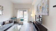 Located at 153-30 89th Avenue in Jamaica, New York, this well-designed residential unit offers a thoughtfully laid-out single-floor living space with a clean, contemporary aesthetic throughout. The open-concept living and dining area serves as the heart of the unit, featuring warm hardwood-style flooring, crisp white walls, and a large window that allows generous natural light to fill the room while offering views of the surrounding neighborhood. The living area provides ample room for a full seating arrangement, and a dedicated media wall with floating shelves adds both function and style. The kitchen is efficiently designed with light wood cabinetry extending to the ceiling for maximum storage, granite countertops, stainless steel appliances including a refrigerator, gas range, and built-in microwave, and a breakfast bar that connects seamlessly to the dining space. The bedroom is a serene retreat, showcasing the same hardwood-style flooring, a bright window dressed with sheer curtains, and clean white walls that create a calm, restful atmosphere. The bathroom is finished with light tile flooring, a wood-toned vanity with an integrated sink, a large mirror, and a shower-tub combination. Throughout the unit, dark-framed doors and trim provide a sharp modern contrast to the light palette, contributing to a cohesive and polished overall design.