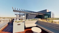 This rooftop terrace is a thoughtfully designed outdoor amenity space that combines modern aesthetics with functional entertaining areas. The deck is finished with rich hardwood planking that adds warmth against the sleek, contemporary design elements throughout. A bold dark-painted pergola structure serves as the centerpiece of the space, providing partial shade while framing sweeping, unobstructed panoramic views of the surrounding cityscape and horizon. Beneath the pergola, a ceiling-mounted outdoor television offers an elevated entertainment experience, complemented by a bar-height counter and seating area nearby. Generous built-in bench seating with clean white finishes lines portions of the deck, creating comfortable lounge zones ideal for gathering. Large planter boxes, planted with ornamental grasses and desert-adapted foliage, are integrated throughout the space, introducing a natural, landscaped character to the rooftop environment. A metal railing perimeter ensures safety while preserving the expansive sightlines to the city skyline visible in multiple directions. The terrace benefits from abundant natural light and open sky exposure, giving the space a bright, airy ambiance throughout the day. Overall, this rooftop terrace delivers a sophisticated, multi-functional outdoor experience with strong architectural detail and striking urban views.