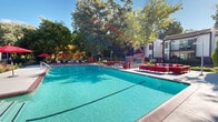Located at 2900 St Paul Dr in Santa Rosa, California, this outdoor amenity area is a well-appointed communal space designed for relaxation and recreation. At its centerpiece sits a generously sized swimming pool with crystal-clear turquoise water, bordered by a classic brick coping and a broad concrete deck. The pool area is thoughtfully furnished with rows of chaise lounges and seating clusters accented by bold red umbrellas, cushions, and planters, creating a vibrant and inviting atmosphere. A built-in brick fireplace feature anchors one end of the deck, providing a cozy gathering spot, while additional outdoor sofas and chairs offer multiple lounging options throughout the space. The entire area is enclosed by a wrought-iron fence with a gated entry, lined with carefully maintained landscaping including ornamental grasses, flowering plants, and mature shade trees that frame the space beautifully against the open California sky. Beyond the pool enclosure, a playground is visible among the greenery, adding a family-friendly element to the community. The surrounding multi-story residential buildings with balconies overlook the pool area, reinforcing the sense of a connected, resort-style community environment. Under clear blue skies, this outdoor space delivers a bright, welcoming, and well-maintained retreat.