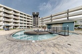 Located in Chattanooga, TN (37408), this expansive outdoor courtyard amenity area is the centerpiece of Chattanooga Flats, a modern multi-story residential community. The courtyard is framed on multiple sides by the building's striking architecture — characterized by rows of private balconies, teal and white exterior accents, and a distinctive glass elevator tower serving as a prominent vertical landmark.

The showpiece of the outdoor space is a generously sized, freeform resort-style swimming pool with vivid turquoise water. A raised island deck positioned at the pool's center provides a unique lounging area outfitted with sculptural white seating, accessible via a bridge walkway with sleek metal railings. Adjacent to the main pool, a separate shallow wading or hot tub area offers a more relaxed aquatic retreat, surrounded by natural stone coping and paver decking.

The surrounding patio is finished with large-format interlocking pavers and offers ample space for outdoor furniture, including lounge chairs and shaded seating areas with umbrellas. A covered patio tucked beneath the building's overhang features vibrant orange Adirondack chairs, adding a pop of color. A manicured green lawn area borders the pool enclosure, and black iron fencing defines the perimeter, creating a cohesive, well-appointed outdoor living environment ideal for relaxation and social gatherings.