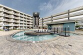 Located in Chattanooga, TN (37408), this expansive outdoor courtyard amenity area is the centerpiece of Chattanooga Flats, a modern multi-story residential community. The courtyard is framed on multiple sides by the building's striking architecture — characterized by rows of private balconies, teal and white exterior accents, and a distinctive glass elevator tower serving as a prominent vertical landmark.

The showpiece of the outdoor space is a generously sized, freeform resort-style swimming pool with vivid turquoise water. A raised island deck positioned at the pool's center provides a unique lounging area outfitted with sculptural white seating, accessible via a bridge walkway with sleek metal railings. Adjacent to the main pool, a separate shallow wading or hot tub area offers a more relaxed aquatic retreat, surrounded by natural stone coping and paver decking.

The surrounding patio is finished with large-format interlocking pavers and offers ample space for outdoor furniture, including lounge chairs and shaded seating areas with umbrellas. A covered patio tucked beneath the building's overhang features vibrant orange Adirondack chairs, adding a pop of color. A manicured green lawn area borders the pool enclosure, and black iron fencing defines the perimeter, creating a cohesive, well-appointed outdoor living environment ideal for relaxation and social gatherings.