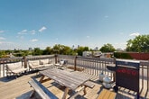 Located in the heart of Montréal, this rooftop space at 4350 Avenue De L'hôtel-De-Ville offers an expansive outdoor terrace with a distinctive dual-level layout. The rooftop is defined by a sturdy wooden boardwalk and railing system that guides visitors across the space, connecting a raised deck area to the broader flat roof surface below. The boardwalk, constructed with weathered wood, adds a warm and functional design element to the otherwise industrial setting.

The lower flat roof section is finished with light-toned concrete and is equipped with several HVAC and mechanical units, reflecting the building's functional infrastructure. A classic red brick facade rises along one side of the rooftop, lending a characteristic Montréal architectural character to the space. Access to the rooftop is provided via a metal door set into a concrete block wall, clearly marked with an emergency exit sign.

The elevated deck area offers panoramic, unobstructed views of the Montreal cityscape, with the downtown skyline visible in the distance and a lush canopy of mature trees framing the mid-ground. The open sky above ensures abundant natural light throughout the day. Whether used as an outdoor gathering space or a communal terrace, this rooftop provides a compelling urban retreat in one of Montréal's vibrant neighborhoods.