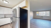 Located at 83 Main St in Newington, Connecticut, this single-floor unit offers a clean, modern interior with thoughtful updates throughout. The open layout is anchored by light-toned vinyl plank flooring that runs consistently across the living areas, creating a cohesive and low-maintenance foundation for the space.

The kitchen is well-appointed with white shaker-style cabinetry extending to the ceiling for maximized storage, granite countertops, a stainless steel refrigerator, and a black electric range with matching stainless accents. A undermount sink with a modern faucet completes the functional workspace. The kitchen opens directly to a spacious living area bathed in natural light from large windows, providing pleasant outdoor views of the surrounding neighborhood.

The living and sleeping areas are generously sized with neutral gray walls, white trim, and continued vinyl plank flooring, offering a versatile canvas for a variety of furniture arrangements. Baseboard heating runs along the perimeter of multiple rooms, ensuring consistent warmth throughout.

The bathroom is finished with large-format gray tile flooring, a white vanity, a soaking tub, and a window that brings in natural light while maintaining privacy. The overall aesthetic throughout the unit is clean, contemporary, and move-in ready.