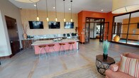 This single-floor community facility is thoughtfully designed to offer a variety of comfortable and functional spaces for gathering, relaxing, and socializing. The grand entrance foyer sets an inviting tone with soaring ceilings, rich wood double doors, and a seamless flow into the main living areas. Crisp white walls and an abundance of natural light from large French doors create a bright, airy atmosphere throughout.

The main lounge area features plush cream sofas, elegant multi-light chandeliers, and wall-mounted flat-screen TVs, providing a comfortable setting for both casual relaxation and group viewing. A dramatic floor-to-ceiling stone fireplace with a rustic wood mantel serves as a stunning focal point, adding warmth and character to the seating area. French doors nearby offer direct access to an outdoor space.

A dedicated bar and counter area showcases a sleek granite-topped island with a salmon-hued base and modern bar stools, complemented by pendant lighting and multiple wall-mounted TVs. This space also features a beverage station, making it ideal for social gatherings. Bold orange accent walls and textured wall finishes add a vibrant, contemporary energy to this zone.

Additional rooms include a dining area and a flexible meeting or activity room with teal accent walls. The consistent use of neutral tones, soft carpeting, and tasteful décor throughout lends the entire space a polished, cohesive character.
