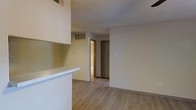This single-floor space features an open-concept layout that connects the kitchen and living areas, creating a cohesive and functional environment. The kitchen includes a white pass-through counter that separates it from the main living area while maintaining an open feel. White cabinetry is visible overhead, contributing to a bright and clean aesthetic throughout.

The flooring consists of light wood-style planks that run consistently across the main living area, adding warmth and a modern touch to the neutral interior. The walls are finished in a soft beige tone, complemented by a textured ceiling that adds subtle visual interest overhead.

A hallway visible from the main living space leads to additional rooms, with two doors providing access to separate areas, likely a bathroom and a closet or bedroom. Ventilation grilles are present both on the ceiling and upper wall, indicating a central HVAC system for climate control.

The space maintains a clean, minimal aesthetic with practical details such as standard electrical outlets and light switches positioned throughout. Overall, the unit presents a straightforward, move-in-ready layout suited for comfortable everyday living.
