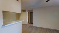 This single-floor space features an open-concept layout that connects the kitchen and living areas, creating a cohesive and functional environment. The kitchen includes a white pass-through counter that separates it from the main living area while maintaining an open feel. White cabinetry is visible overhead, contributing to a bright and clean aesthetic throughout.

The flooring consists of light wood-style planks that run consistently across the main living area, adding warmth and a modern touch to the neutral interior. The walls are finished in a soft beige tone, complemented by a textured ceiling that adds subtle visual interest overhead.

A hallway visible from the main living space leads to additional rooms, with two doors providing access to separate areas, likely a bathroom and a closet or bedroom. Ventilation grilles are present both on the ceiling and upper wall, indicating a central HVAC system for climate control.

The space maintains a clean, minimal aesthetic with practical details such as standard electrical outlets and light switches positioned throughout. Overall, the unit presents a straightforward, move-in-ready layout suited for comfortable everyday living.