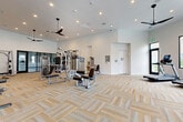 Located at 12406 Clark St in Moreno Valley, CA, this well-appointed fitness center offers a generously sized, single-floor workout facility designed to accommodate a full range of exercise needs. The open layout provides ample floor space, allowing for comfortable movement between equipment zones without congestion.

The interior features a clean, modern aesthetic with crisp white walls, elevated ceilings, and recessed lighting complemented by stylish pendant ceiling fans that ensure consistent airflow throughout the space. The floor is finished with a textured, neutral-toned carpet tile in a herringbone-style pattern, adding visual warmth while providing cushioning underfoot.

The strength training area is well-equipped with cable machines, a multi-station weight machine, a Smith machine, a flat bench, and a dedicated dumbbell rack with a full set of free weights—all positioned alongside large wall mirrors that enhance the sense of space. A functional training wall features slatted storage for medicine balls, kettlebells, battle ropes, and a punching bag, supporting a variety of workout styles.

The cardio zone includes treadmills and elliptical machines, positioned near large black-framed windows and glass doors that invite natural light and provide outdoor views. A water fountain is also accessible within the space. The facility's layout and equipment selection make it a comprehensive, professional-grade fitness destination.