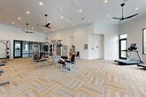 Located at 12406 Clark St in Moreno Valley, CA, this well-appointed fitness center offers a generously sized, single-floor workout facility designed to accommodate a full range of exercise needs. The open layout provides ample floor space, allowing for comfortable movement between equipment zones without congestion.

The interior features a clean, modern aesthetic with crisp white walls, elevated ceilings, and recessed lighting complemented by stylish pendant ceiling fans that ensure consistent airflow throughout the space. The floor is finished with a textured, neutral-toned carpet tile in a herringbone-style pattern, adding visual warmth while providing cushioning underfoot.

The strength training area is well-equipped with cable machines, a multi-station weight machine, a Smith machine, a flat bench, and a dedicated dumbbell rack with a full set of free weights—all positioned alongside large wall mirrors that enhance the sense of space. A functional training wall features slatted storage for medicine balls, kettlebells, battle ropes, and a punching bag, supporting a variety of workout styles.

The cardio zone includes treadmills and elliptical machines, positioned near large black-framed windows and glass doors that invite natural light and provide outdoor views. A water fountain is also accessible within the space. The facility's layout and equipment selection make it a comprehensive, professional-grade fitness destination.