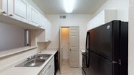 This single-floor residence offers a thoughtfully organized layout that balances functionality and comfort across its key living areas. The galley-style kitchen is a standout feature, lined on both sides with crisp white cabinetry that provides generous storage from floor to ceiling. Light-toned countertops, a double stainless steel sink, and black appliances — including a refrigerator and range — complement the space, while light wood-style flooring ties the room together. A door at the end of the kitchen leads to a dedicated laundry area, adding everyday convenience. The living room is warm and inviting, featuring light hardwood-style flooring, a ceiling fan with light fixture, and multiple windows including a glass-paneled exterior door that floods the room with natural light and offers a view of lush greenery outside. The bedroom is carpeted for comfort, equipped with its own ceiling fan, a window for natural light, and direct access to the living area. The unit includes two bathrooms: one featuring a vanity with a large mirror and single basin sink, and a second full bathroom complete with a bathtub/shower combo, toilet, and a white vanity with ample counter space. Neutral tones throughout the space create a clean, versatile backdrop ready for personalization.
