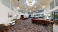 Located at 928 Shorepoint Ct in Alameda, CA, this single-floor clubhouse facility is a thoughtfully designed community gathering space that blends coastal charm with modern sophistication. The centerpiece of the space is a dramatic atrium-style great room crowned by a soaring geometric vaulted ceiling with a central skylight, allowing abundant natural light to pour in. Suspended globe pendant lights complement the architectural detail above, while the open floor plan accommodates multiple distinct seating arrangements—including mid-century modern lounge chairs, a large sectional sofa, and intimate conversation nooks.

A secondary lounge area features a built-in curved banquette with plush cushions, exposed ductwork overhead lending an industrial touch, and direct access to an outdoor deck. The dining room is furnished with a long communal table and Scandinavian-style woven chairs, illuminated by striking birdcage pendant lights, and opens to the exterior via floor-to-ceiling glass doors framed by soft linen drapes.

The kitchen area is equipped with a large center island with bar seating, warm wood cabinetry, white quartz countertops, and modern pendant lighting. A utility nook with an undermount sink and light wood cabinetry provides additional functionality. Throughout, white shiplap walls, rich hardwood and patterned carpet flooring, and curated artwork create a cohesive, welcoming ambiance.