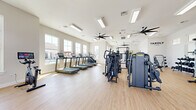 The Yardly Fitness Center at Avalon Landing in Cypress, Texas is a thoughtfully designed, single-floor community fitness facility that offers a comprehensive range of exercise options in a bright, welcoming environment. The space features light hardwood-style flooring throughout, crisp white walls, and an abundance of natural light streaming in through large windows that frame views of the surrounding neighborhood.

The main workout floor is generously proportioned and organized into distinct training zones. A dedicated cardio section lines the windowed wall with multiple treadmills, elliptical cross-trainers, stationary cycling bikes, and rowing machines — all equipped with digital displays. A strength training area hosts a robust selection of cable and multi-function machines, ideal for resistance-based workouts. A separate functional training zone includes a well-stocked equipment rack with kettlebells, medicine balls, resistance accessories, and a wall-mounted pad for dynamic exercises.

Large ceiling fans are distributed throughout the space, ensuring consistent airflow and comfort during workouts. Overhead linear lighting supplements the ample natural light. A mirrored wall section enhances the sense of openness, while the Yardly branding adds a polished, professional aesthetic. Glass double doors provide easy access to the outdoor area, where a pool is visible, seamlessly connecting indoor fitness with outdoor amenities.