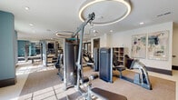 This fitness center offers a comprehensive workout environment thoughtfully designed with both aesthetics and functionality in mind. The space is organized into distinct training zones, beginning with a strength and machine area outfitted with TRUE-brand cable systems, weight machines, and adjustable benches. A dedicated free weights section features a full dumbbell rack displayed against a floor-to-ceiling mirror wall, allowing users to monitor their form during exercise. A separate functional training zone is anchored by a wall-mounted multi-purpose rig stocked with medicine balls, kettlebells, resistance bands, TRX straps, and a plyo box, set against a bold teal accent wall that adds a vibrant design element to the space.

The facility is finished with neutral-toned carpet tiles and hard flooring, offering durable and practical surfaces throughout. Overhead, a series of distinctive circular LED light fixtures serve as both a stylish focal point and a source of consistent, even illumination. Cubbies and shelving provide storage for personal items and accessories. Abstract artwork adorns the white walls, softening the industrial feel of the equipment. Large windows along one wall invite natural light and outdoor views, contributing to an energizing and welcoming atmosphere.