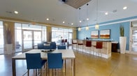 This single-floor clubhouse offers a bright, open-concept gathering space designed for comfort and socializing. The interior is characterized by soaring ceilings with a distinctive wood-paneled ceiling accent, recessed lighting, and elegant pendant lights that hang above the bar counter, creating a warm and inviting ambiance.

The space features a built-in bar area finished in light wood, equipped with a countertop workspace and what appears to be a coffee station, complemented by rich dark wood barstools with chrome legs. Framed artwork adorns the blue accent wall behind the bar, adding a refined aesthetic touch.

Throughout the main area, white rectangular tables are paired with navy blue upholstered chairs, offering flexible seating arrangements suitable for casual gatherings or meetings. The polished concrete flooring in warm amber tones ties the space together seamlessly.

Large floor-to-ceiling windows and glass entry doors along the front facade flood the interior with abundant natural light while providing open views to the exterior. The color palette — combining sky blue accent walls, cream tones, and warm wood elements — creates a contemporary yet welcoming atmosphere. Overall, this clubhouse is thoughtfully designed to serve as a versatile and stylish communal hub.