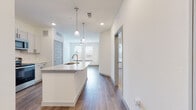 This single-floor unit is designed with a clean, contemporary aesthetic throughout. The open-concept layout connects the kitchen and living area seamlessly, creating a bright and functional living space. Crisp white walls, recessed lighting, and wood-style plank flooring run consistently through the main areas, contributing to a cohesive and modern feel.

The kitchen is well-appointed, featuring gray shaker-style cabinetry, a granite-topped island with an integrated sink, and a full suite of stainless steel appliances including a refrigerator, range, and microwave. Pendant lights hang above the island, adding both task lighting and visual interest. The kitchen flows directly into the living area, which benefits from natural light provided by windows at the far end of the space.

The bathroom includes a full soaking tub and shower combination with vertically tiled surround, wood-look flooring, a white vanity with granite countertop, and matte black hardware accents. A dedicated laundry area houses a full-size top-load washer and dryer alongside built-in shelving for storage. Distinctive louvered interior doors are featured throughout, adding a subtle architectural detail that complements the unit's modern design.
