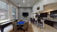 This single-floor game room in Humble, TX is thoughtfully designed for entertainment and social activity. The main area is anchored by a dedicated poker table with comfortable upholstered seating, complemented by a separate high-top bar table with leather barstools — perfect for casual dining or card games. Soaring ceilings and floor-to-ceiling windows flood the space with natural light while offering views of lush outdoor greenery, creating an open and inviting atmosphere. Neutral gray walls, recessed lighting, and large-format tile flooring throughout lend a clean, modern aesthetic. Built-in display shelves add architectural interest and subtle decorative accents. The kitchenette is fully equipped, featuring rich dark cabinetry, a mosaic tile backsplash, granite countertops, stainless steel appliances including a microwave and dishwasher, and a dedicated coffee station. A curved granite-topped bar with pendant lighting serves as a seamless transition between the kitchen and the main gathering area. French doors connect the game room to an adjacent lounge space, enhancing the flow for larger events. This versatile space is well-suited for community use, private gatherings, or recreational activities.