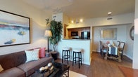 This single-floor residence offers a thoughtfully designed open-concept layout that seamlessly connects the living, dining, and kitchen areas. The living room is a welcoming space featuring warm hardwood-style flooring, large windows that invite generous natural light, and ample room for comfortable seating arrangements. A ceiling fan overhead and neutral-toned walls create a relaxed, airy atmosphere. Framed artwork and a glass-top coffee table add refined touches to the space.

The kitchen is positioned behind a half-wall peninsula with granite-style countertops and two bar stools, making it ideal for casual dining. Inside, warm wood cabinetry pairs with black appliances, including a refrigerator, range, and built-in microwave, all set above durable wood-style flooring. Track lighting illuminates the workspace effectively.

The bedrooms are carpeted for comfort, each featuring a ceiling fan, natural light from windows dressed with fabric valances and blinds, and access to closet storage. The rooms offer a calm, restful ambiance with neutral walls and tasteful furnishings.

The bathroom features rich wood cabinetry, granite-style countertops, a tub-shower combination, and hardwood-style flooring, delivering a clean and functional design throughout.