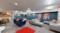 Located at 313 SW 13th St in Gainesville, Florida, this single-floor amenity lounge offers a generously proportioned, open-plan recreation and social space. The room is designed with a contemporary aesthetic, combining bold teal and warm terracotta accent walls with neutral white surfaces and textured tile-pattern carpeting throughout. Vivid red area rugs punctuate the floor plan, adding energy and definition to distinct zones within the space.

The lounge is organized into several functional areas. A game zone anchors the center of the room, featuring a full-size ping-pong table as its focal point, complemented by a wall-mounted flat-screen television. A comfortable seating area includes a tufted navy blue velvet sofa and a large gray sectional, arranged to encourage gathering and relaxation. Bold, colorful pop-art pieces adorn the teal accent wall, contributing to the room's lively atmosphere.

A dining area with a solid wood table and vibrant yellow chairs provides space for group meals or activities, accented by original artwork nearby. A compact kitchenette with white cabinetry, a granite countertop, open shelving with greenery, and a small appliance station rounds out the space. Recessed and overhead fluorescent lighting ensures the entire area is well illuminated, creating an inviting, energetic environment suited for community engagement.