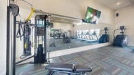 Located at 19201–19209 Kuykendahl Rd in Houston, TX, this single-floor fitness center is designed to provide a comprehensive and comfortable workout environment. The space is defined by impressively high ceilings accented with exposed decorative beams, creating an open and airy atmosphere throughout. Multiple ceiling fans and recessed lighting ensure consistent airflow and illumination across the floor.

Natural light pours in through a series of tall windows and clerestory windows positioned along the upper walls, brightening the interior and offering views of the surrounding greenery. Large wall-to-wall mirrors are strategically placed to enhance the sense of space and support proper exercise form.

The facility is well-equipped with a wide variety of cardio machines, including treadmills and ellipticals, as well as a broad selection of strength-training equipment such as cable machines, multi-station weight machines, and benches. A dedicated free weights area features a full rack of dumbbells in graduated sizes, complemented by kettlebells and fitness accessories including yoga and foam rollers.

The flooring consists of multi-toned carpet tiles in teal, blue, and neutral tones, adding both comfort underfoot and visual character. Wall-mounted televisions are positioned for easy viewing during workouts. The overall design balances functionality with an inviting, modern aesthetic suited for a wide range of fitness activities.