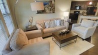This single-floor space offers a welcoming, open-concept layout that seamlessly connects the living area to the kitchen. The living room is thoughtfully arranged with a plush, neutral-toned sofa and matching accent chair, centered around a robust metal-framed coffee table with a dark, industrial-style surface. Decorative throw pillows in geometric patterns add visual interest, while ambient floor and table lamps create a warm, inviting atmosphere throughout the space.

A colorful Mediterranean-inspired painting adorns the wall above the sofa, serving as a focal point that adds character to the room. End tables with elegant accessories, including a sculptural clock and a crystal-based lamp, complement the refined yet approachable aesthetic.

The kitchen, visible in the background, features dark cabinetry with black appliances, offering a striking contrast to the lighter tones of the living area. Light-colored flooring runs throughout, unifying the open layout and enhancing the sense of spaciousness. Overall, the space strikes a balance between comfort and style, with neutral tones and carefully chosen décor creating a cohesive and livable environment.