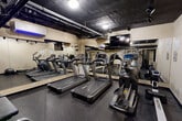 Located at 1530 16th St NW in Washington, DC, this fitness center is a well-equipped exercise room designed to support a comprehensive workout routine. The space features an exposed, dark-painted ceiling with visible ductwork and overhead track lighting, lending it an industrial aesthetic that keeps the atmosphere focused and energized. Rubber flooring throughout provides both safety and durability underfoot.

The cardio section is anchored by multiple treadmills and a stair climber, all positioned along a wall of floor-to-ceiling mirrors that visually expand the room and allow users to monitor their form. A wall-mounted flat-screen television is installed above the mirrors, offering entertainment during cardio sessions. A split-system air conditioning unit ensures climate comfort year-round.

On the strength-training side, the room houses a dual-cable Cybex functional trainer, a rack of hex dumbbells in varying weights, a stability ball, and a flat workout bench, providing a solid range of resistance training options. A sanitizing station is conveniently positioned near the exit, promoting cleanliness. The neutral beige walls keep the space feeling clean and open. The room connects directly to a hallway with decorative tile flooring, integrating seamlessly into the larger building amenity area.