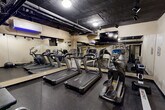 Located at 1530 16th St NW in Washington, DC, this fitness center is a well-equipped exercise room designed to support a comprehensive workout routine. The space features an exposed, dark-painted ceiling with visible ductwork and overhead track lighting, lending it an industrial aesthetic that keeps the atmosphere focused and energized. Rubber flooring throughout provides both safety and durability underfoot.

The cardio section is anchored by multiple treadmills and a stair climber, all positioned along a wall of floor-to-ceiling mirrors that visually expand the room and allow users to monitor their form. A wall-mounted flat-screen television is installed above the mirrors, offering entertainment during cardio sessions. A split-system air conditioning unit ensures climate comfort year-round.

On the strength-training side, the room houses a dual-cable Cybex functional trainer, a rack of hex dumbbells in varying weights, a stability ball, and a flat workout bench, providing a solid range of resistance training options. A sanitizing station is conveniently positioned near the exit, promoting cleanliness. The neutral beige walls keep the space feeling clean and open. The room connects directly to a hallway with decorative tile flooring, integrating seamlessly into the larger building amenity area.