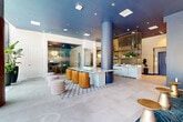 This sophisticated single-floor lobby and entrance space makes a striking first impression through its bold contemporary design and carefully curated aesthetic. Large floor-to-ceiling windows at the entrance flood the space with natural light while offering open views of the surrounding streetscape, creating a welcoming and airy atmosphere.

The entrance area features large-format porcelain tile flooring that transitions into a geometric hexagonal patterned tile, clearly defining functional zones throughout the space. Structural columns finished in deep navy blue anchor the open layout and complement the dramatic painted ceiling, which serves as a standout architectural feature. A central reception or concierge counter with a marble-topped surface and warm wood barrel stools provides a natural focal point.

A comfortable lounge area is appointed with a plush teal sectional sofa, vibrant area rugs, and an eye-catching large-scale art piece adorning the wall. Elegant globe pendant chandeliers cast a warm, ambient glow across the seating areas. Glass-walled private rooms with gold-accented pendant lighting are accessible directly from the main floor, offering intimate meeting or office spaces without sacrificing visual openness. A built-in shelving display unit with gold metal slat detailing adds a refined, boutique-hotel quality to the overall design. Throughout, a cohesive palette of navy, teal, gold, and warm wood tones creates a polished and vibrant ambiance.