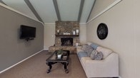 This single-story community clubhouse is designed to serve as a versatile gathering space, combining comfort and functionality throughout its open layout. The architecture is defined by vaulted ceilings with exposed painted wood beams, which lend a warm, rustic character to the interior while adding a sense of height and openness.

The lounge area serves as the heart of the space, anchored by a striking floor-to-ceiling stone fireplace with an ornate iron screen and a wooden mantel. Plush neutral-toned seating and a glass-top coffee table furnish the area, while a wall-mounted television adds a modern convenience. Tile flooring surrounds the hearth, transitioning to carpeting throughout the remainder of the space.

Adjacent to the lounge is a dedicated Wi-Fi bar — a built-in workstation with seating for multiple users, clearly labeled and equipped with devices for resident use. This area reflects the space's commitment to practical amenities.

The dining area occupies a bright, airy section of the clubhouse lined with large windows that flood the room with natural light and offer views of the surrounding outdoor greenery. Multiple high-top and standard dining tables with dark wood chairs provide ample seating for gatherings. A secondary entrance with glass-paneled doors connects this area directly to the exterior.
