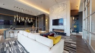 Located in Denver's Lowry neighborhood, this upscale communal lounge space exemplifies contemporary design with a carefully curated aesthetic. The main gathering area is anchored by a striking floor-to-ceiling marble accent wall housing a linear gas fireplace, creating a bold focal point complemented by a wall-mounted flat-screen TV. Rich veining in the marble adds natural texture and drama to the space.

The lounge features an expansive seating arrangement with a large, plush sectional sofa accented by jewel-toned throw pillows in gold and burgundy, all set atop a multi-toned striped area rug. Recessed and cove lighting, along with cascading pendant lights near the bar area, create a warm, layered ambiance throughout.

A wet bar and lounge bar area with dark tile, warm wood shelving, and counter-height seating provides a dedicated social hub. Adjacent to this, floor-to-ceiling glass curtain walls flood the dining nook with abundant natural light, offering sweeping panoramic views of the Denver skyline and Rocky Mountains beyond. A round dining table with cognac leather chairs makes this a refined spot for casual gatherings.

Additional areas feature wide-plank flooring, dark accent walls, and natural greenery accents, reinforcing the space's modern yet inviting character throughout.