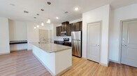 This single-floor residence showcases a thoughtfully designed open-concept layout, seamlessly connecting the kitchen and living areas for an airy, functional feel. The kitchen serves as the centerpiece of the space, anchored by a large island topped with striking granite countertops and fitted with a double stainless steel undermount sink and a pull-down faucet. Dark espresso cabinetry provides generous storage and creates a bold contrast against the crisp white walls and light wood-look flooring that runs throughout. A full suite of stainless steel appliances — including a refrigerator, range, and microwave — completes the well-appointed cooking space. Elegant pendant lights hang above the island, adding warmth and a contemporary touch. The living area benefits from natural light flowing in through windows and glass French doors, creating a bright and welcoming atmosphere. An arched entryway adds a subtle architectural detail to the open floor plan. The bathroom features a dual-sink granite vanity with dark cabinetry, a large frameless mirror, and a tiled shower, maintaining the cohesive modern aesthetic found throughout the residence. Light-toned luxury vinyl flooring, recessed lighting, and clean white walls unify the space with a fresh, move-in-ready appeal.