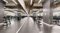 This large-scale fitness facility offers a comprehensive, single-floor exercise environment designed to accommodate a wide range of workout disciplines. The space features an industrial-modern aesthetic, with exposed concrete ceilings, sleek linear LED lighting, and durable speckled rubber flooring throughout the main exercise areas.

The cardio zone is lined with a row of treadmills and other aerobic machines, all positioned alongside floor-to-ceiling mirrors that enhance the sense of openness. Adjacent to this area, a dedicated strength training zone features multiple power racks, a full complement of dumbbells arranged on angled racks, adjustable benches, and a broad selection of resistance machines suited for both upper and lower body training.

A separate functional training area provides open floor space alongside kettlebells, medicine balls, stability equipment, and wall-mounted accessories for versatile, freeform workouts. The facility also includes a bright, dedicated studio room with light wood flooring, mirrored walls, and ample open space outfitted with boxing dummies and resistance training attachments — ideal for group classes or martial arts training.

The entrance to the main floor is framed by frosted glass doors with modern hardware, lending a polished, professional appearance. Exposed ductwork and structural columns add an urban industrial character, while the thoughtful layout ensures clear circulation throughout the entire facility.