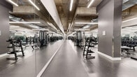This large-scale fitness facility offers a comprehensive, single-floor exercise environment designed to accommodate a wide range of workout disciplines. The space features an industrial-modern aesthetic, with exposed concrete ceilings, sleek linear LED lighting, and durable speckled rubber flooring throughout the main exercise areas.

The cardio zone is lined with a row of treadmills and other aerobic machines, all positioned alongside floor-to-ceiling mirrors that enhance the sense of openness. Adjacent to this area, a dedicated strength training zone features multiple power racks, a full complement of dumbbells arranged on angled racks, adjustable benches, and a broad selection of resistance machines suited for both upper and lower body training.

A separate functional training area provides open floor space alongside kettlebells, medicine balls, stability equipment, and wall-mounted accessories for versatile, freeform workouts. The facility also includes a bright, dedicated studio room with light wood flooring, mirrored walls, and ample open space outfitted with boxing dummies and resistance training attachments — ideal for group classes or martial arts training.

The entrance to the main floor is framed by frosted glass doors with modern hardware, lending a polished, professional appearance. Exposed ductwork and structural columns add an urban industrial character, while the thoughtful layout ensures clear circulation throughout the entire facility.