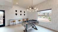Located at 9600 W Georgia Ave in Glendale, Arizona, this single-floor game room offers a generously sized recreational space designed with a contemporary aesthetic. The room is finished with large-format neutral tile flooring that enhances the bright, open feel of the interior. A striking sputnik-style chandelier with exposed Edison bulbs serves as a bold focal point, casting warm ambient light throughout the space.

The game room is anchored by a full-size foosball table and complemented by a wall-mounted shuffleboard table, providing a range of entertainment options. A built-in counter-height ledge with cork-top bar stools lines one wall, offering casual seating beneath an eye-catching arrangement of sculptural black mask wall art. Framed black-and-white artwork decorates an adjacent wall, adding an artistic touch to the playful environment.

Large windows and a glass-paneled door flood the space with natural light while framing views of the surrounding outdoor areas. The room flows openly into an adjacent lounge area, which features sophisticated furnishings, a wood-accented ceiling detail, a fireplace with backlit shelving, and additional seating — creating a seamless transition between active recreation and relaxed socializing. Direct outdoor access further extends the usability of this well-appointed amenity space.