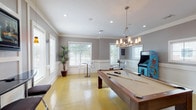 This single-story recreational facility in Humble, TX offers a well-appointed community space designed for both leisure and fitness. The game room serves as a central hub, featuring a full-size pool table beneath an elegant multi-light chandelier, a classic arcade cabinet, and a Coca-Cola vending machine for refreshments. The polished concrete floors in a warm golden hue extend throughout, providing a cohesive aesthetic across all areas. Crown molding and white wainscoting add a refined architectural touch to the gray-toned walls, while multiple windows fitted with white blinds allow natural light to fill the space. A dedicated lounge area along one wall includes a wall-mounted flat-screen television, a sleek bar-height counter with padded stools, and accent pendant lighting. Transparent bistro tables and chairs are positioned near the windows, offering casual seating throughout. An adjacent fitness center, visible through large interior glass windows and accessible via a white-paneled door, houses cardio equipment including treadmills and stationary bikes, as well as strength-training machines, all beneath ceiling fans that promote airflow during workouts.
