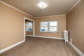 This single-floor unit offers a practical layout comprising two bedrooms, two bathrooms, a kitchen, a hallway, and a laundry area. The primary bedroom is notably spacious relative to the overall footprint, featuring wall-to-wall carpeting, crown molding, and multiple windows that allow natural light to fill the room. A vintage cast-iron radiator provides a classic architectural detail. Rich, dark-stained wooden doors add a traditional character throughout. A closet accessible from the primary bedroom provides dedicated storage space.

The second bedroom also features carpeting, crown molding, and windows with outdoor views, as well as access to a refrigerator, suggesting a functional secondary living area. The kitchen is equipped with oak cabinetry, dark laminate countertops, a gas range, and a single-basin stainless steel sink, all set on stone-tile flooring.

Both bathrooms are finished with neutral stone-tile flooring and include a toilet, pedestal-style vanity, and mirrored medicine cabinet. The hallway connecting the rooms features matching tile flooring and built-in drawer storage. A narrow corridor with hard flooring serves as an additional storage or laundry space, completing the efficient and functional layout of this unit.