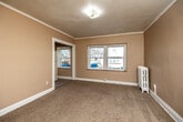 This single-floor unit offers a practical layout comprising two bedrooms, two bathrooms, a kitchen, a hallway, and a laundry area. The primary bedroom is notably spacious relative to the overall footprint, featuring wall-to-wall carpeting, crown molding, and multiple windows that allow natural light to fill the room. A vintage cast-iron radiator provides a classic architectural detail. Rich, dark-stained wooden doors add a traditional character throughout. A closet accessible from the primary bedroom provides dedicated storage space.

The second bedroom also features carpeting, crown molding, and windows with outdoor views, as well as access to a refrigerator, suggesting a functional secondary living area. The kitchen is equipped with oak cabinetry, dark laminate countertops, a gas range, and a single-basin stainless steel sink, all set on stone-tile flooring.

Both bathrooms are finished with neutral stone-tile flooring and include a toilet, pedestal-style vanity, and mirrored medicine cabinet. The hallway connecting the rooms features matching tile flooring and built-in drawer storage. A narrow corridor with hard flooring serves as an additional storage or laundry space, completing the efficient and functional layout of this unit.