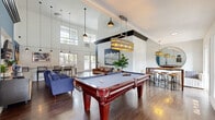 This thoughtfully designed single-floor communal space functions as a multifaceted lounge and recreation area, offering a generous open-plan layout that feels remarkably spacious thanks to its soaring vaulted ceilings accented by bold dark exposed beams. Rich hardwood flooring runs throughout, anchoring the room's eclectic yet cohesive aesthetic. At the heart of the space sits a striking cherry-wood pool table, illuminated by a large drum pendant light with gold-toned accents, making it a natural focal point for recreation and socializing. Adjacent to the game area, a comfortable lounge zone features vibrant cobalt-blue sofas and armchairs arranged around a glass coffee table, set against a dramatic teal-blue feature wall that houses a wall-mounted flat-screen TV. A sleek wet bar with a dark island, counter seating, and pendant lighting provides a functional refreshment station, while a built-in banquette and high-top dining table with industrial stools offer a casual dining nook accented by patterned wallpaper and a large round mirror. Expansive gridded windows and glass-panel doors flood the space with natural light and offer views of the surrounding outdoor environment, enhancing the open and inviting ambiance of this well-appointed communal gathering space.